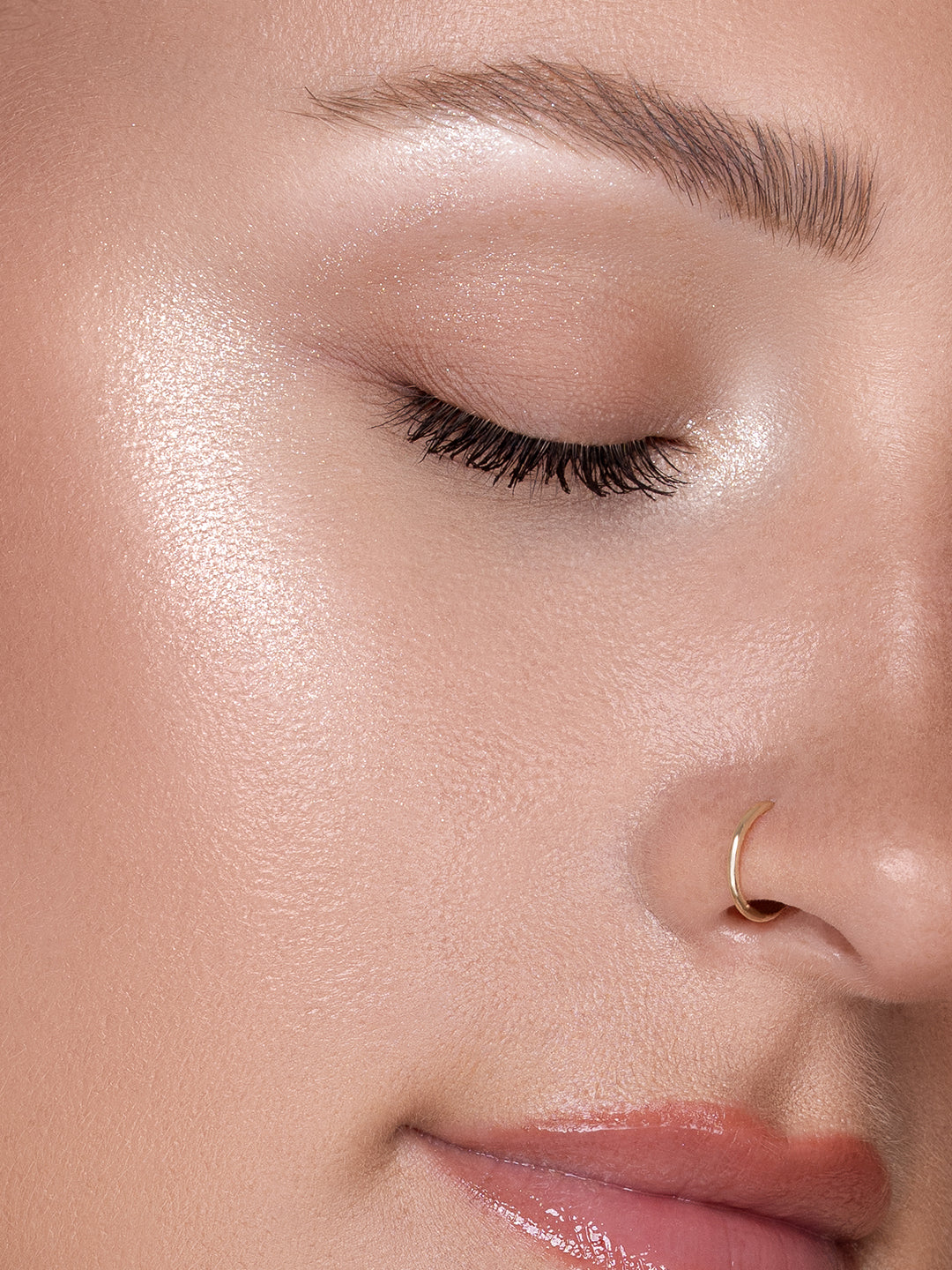Close-up of a woman's eye with a subtle makeup look and gold nose ring Shimmer Veil highlighter by Beauty Creations