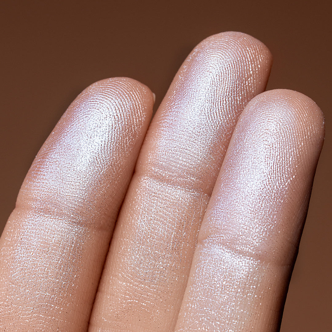 Close-up of a hand with a brown background showing a swatch of Shimmer Veil face and body highlighter by Beauty Creations. Color payoff.