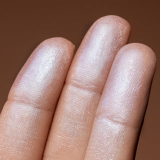 Close-up of a hand with a brown background showing a swatch of Shimmer Veil face and body highlighter by Beauty Creations. Color payoff.