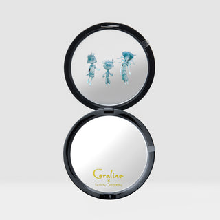 An open round compact mirror with a black frame, showing a reflective silver interior. The top half contains a printed illustration of three small blue cartoon characters, and the bottom half has the brand name 'Coraline x Beauty Creations' printed in gold near the bottom edge.