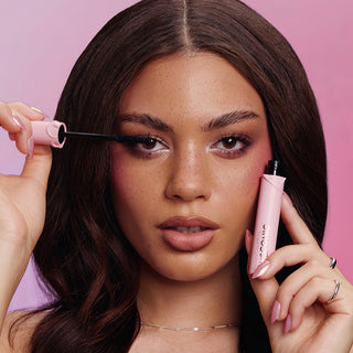 Woman applying mascara with a pink mascara wand against a pink background