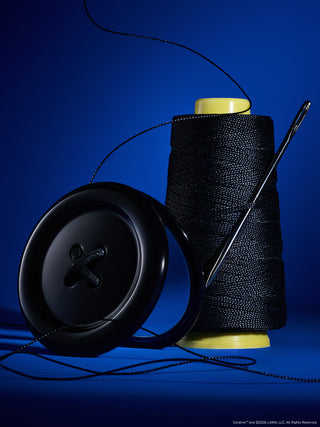 A studio product shot featuring a large black button, a spool of black thread with yellow plastic ends, and a metal needle on a deep blue background.