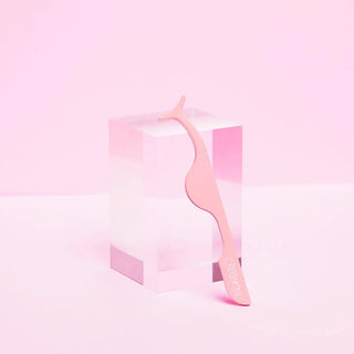 A pink Beauty Creations ergonomic eyelash applicator resting against a clear acrylic block on a pastel pink background.